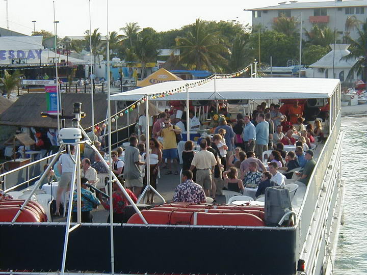 Crowd of people socializing with drinks on the canopied upper deck of a party boat docked at a marina at golden hour. String lights hang overhead; waterfront shops, palm trees and a 'TUESDAY' sign are visible behind.