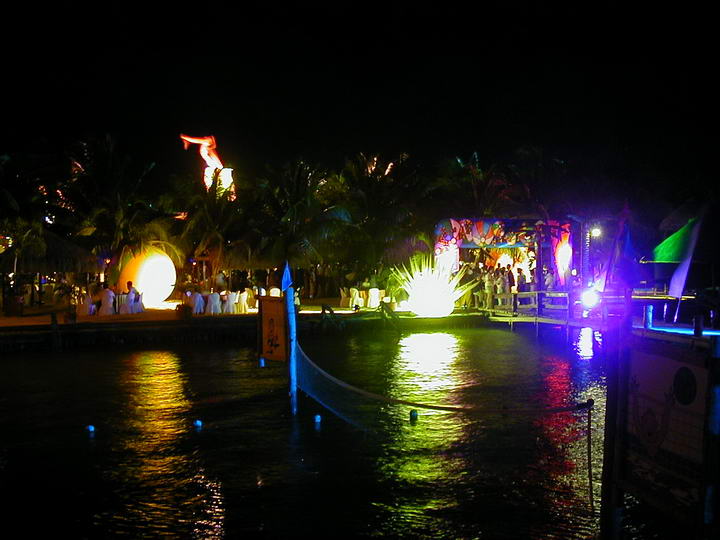 Nighttime waterfront party scene viewed across dark water, with colorful stage lighting, fire/flame effects, illuminated palm trees and crowds of people dressed in white along the shore. Light reflections shimmer on the water.