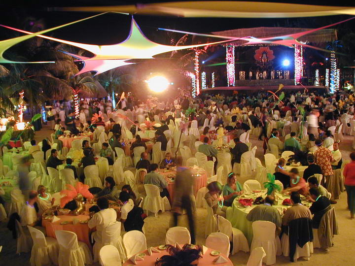 Elevated view of a large nighttime beach banquet with hundreds of guests at round tables dressed in white, peach and green linens, colorful shade sails overhead, a lit stage and palm trees. Glow sticks and feathers add a festive party mood.