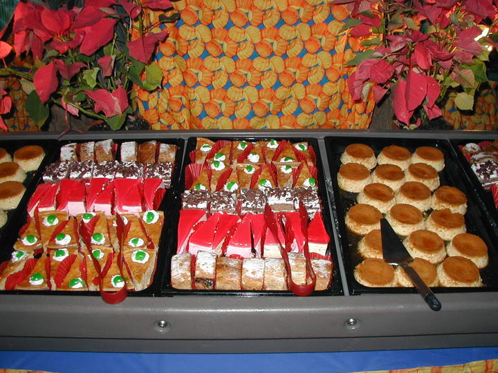 Close-up of a dessert buffet with trays of assorted pastries, layered cakes with whipped cream, strawberry-topped slices and round biscuits, garnished with red poinsettia flowers and an orange patterned cloth backdrop.