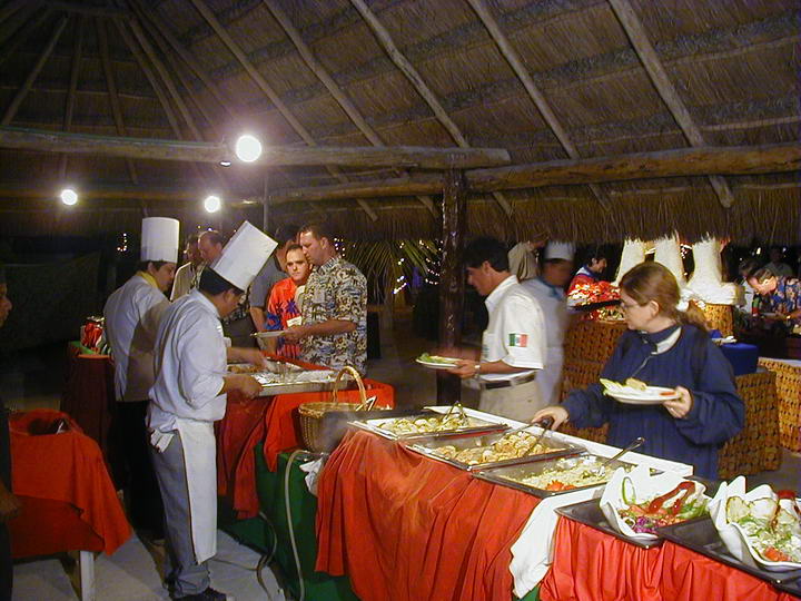 Buffet line under a thatched palapa roof where uniformed chefs in white toques serve guests from chafing dishes draped in red and orange cloth. Several guests hold plates of food; string lights twinkle in the background.