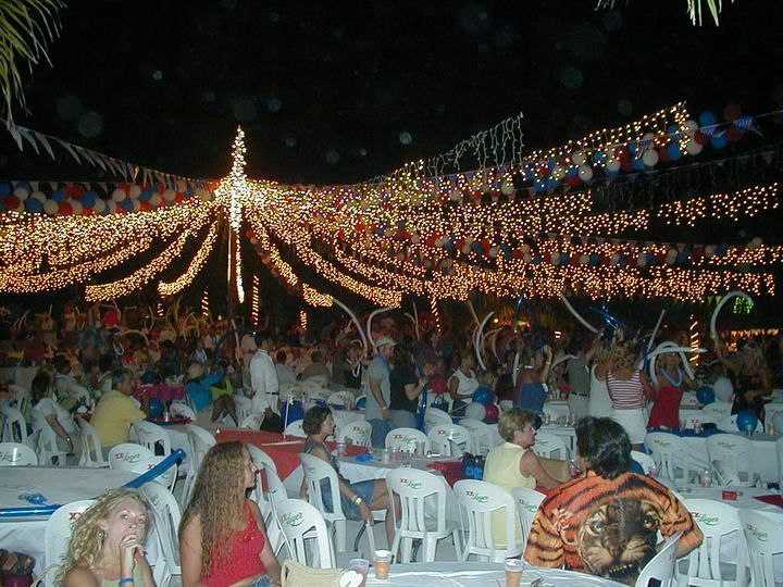 Crowded nighttime beach party under a canopy of thousands of warm fairy lights and balloons, with many guests seated at white-clothed round tables. People hold inflatable noise sticks; the festive atmosphere is dense and lively.