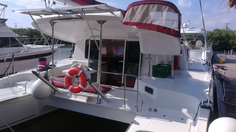 Stern and cockpit view of a white catamaran with a red canopy and an orange life ring at a marina, the salon visible through the doorway, other boats nearby, sunny.