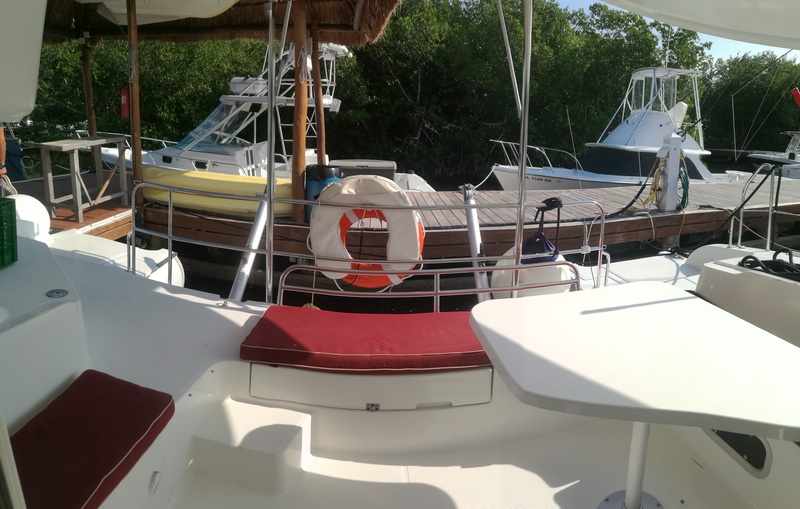Cockpit seating of a catamaran with red cushions and a white life ring, looking toward mangroves and sportfishing boats at a marina, sunny.