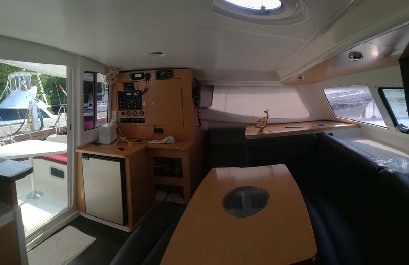 Interior salon and nav station of a catamaran with light wood finish, an instrument panel, a refrigerator, a settee and a table with a circular inset, doors opening to the cockpit, bright daylight.