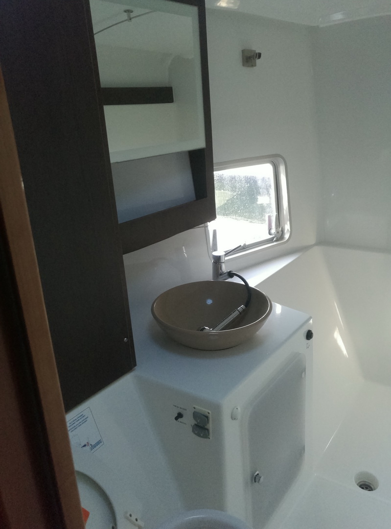 Interior head/bathroom of a catamaran with a white sink basin, a mirrored cabinet, a small window and dark wood trim.