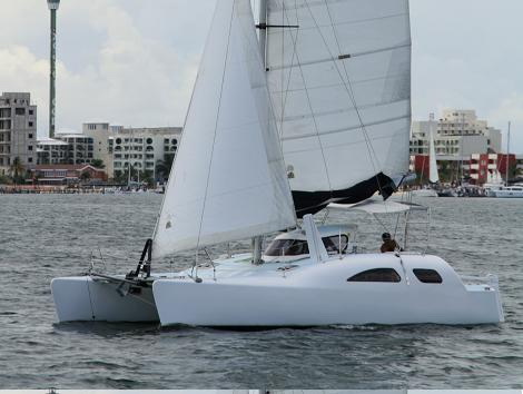 Sleek white performance sailing catamaran with sails up, gliding on calm grey water, a single person aboard. City buildings line the shore behind under an overcast sky.
