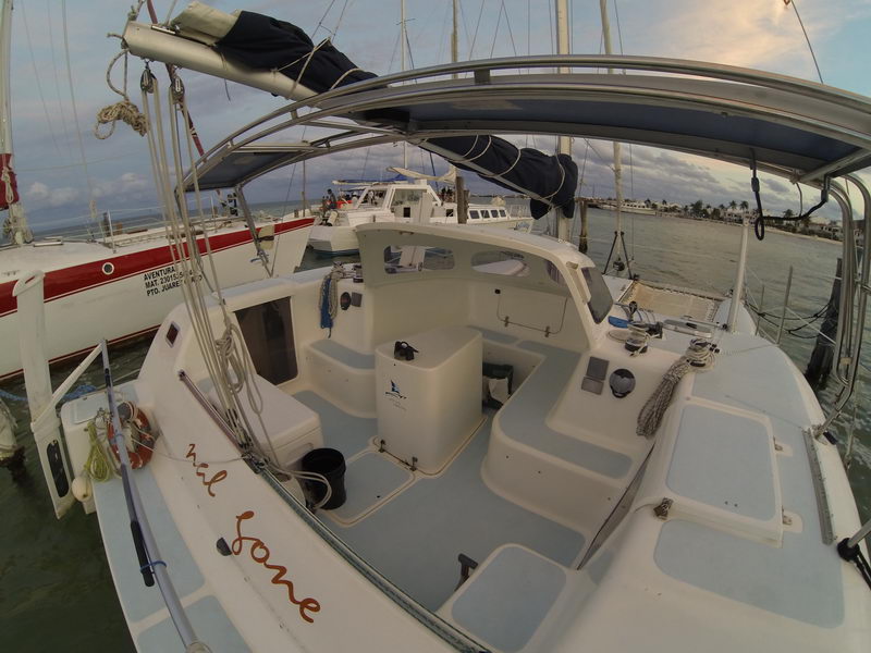 Wide-angle (GoPro) cockpit view of the sailing catamaran 'Nai Lone' at a marina at dusk, white cockpit with the helm console and the name in script on the transom. Other sailboats moored alongside under a soft sunset sky.