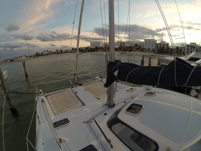 Wide-angle foredeck view of a catamaran at the marina at dusk, trampoline nets and deck hatches in the foreground, calm water with waterfront buildings and palm trees in the soft evening light.
