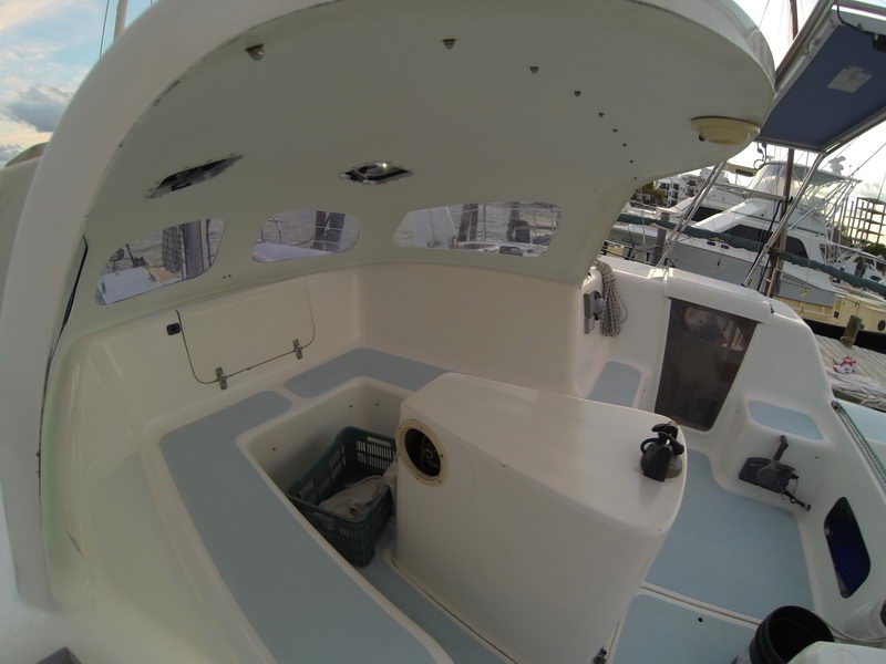 Wide-angle view of a catamaran's cockpit interior, white fiberglass seating and a helm pod with a basket of items/laundry. Marina and other boats visible in the evening light.