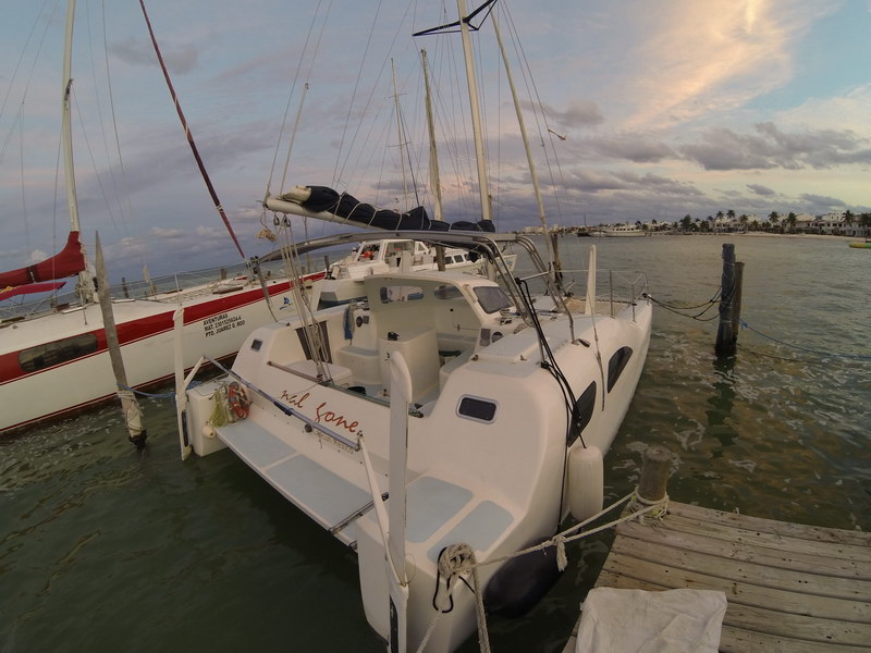 Wide-angle stern view of the 'Nai Lone' sailing catamaran moored at a dock at dusk, the name in script on the hull, a red multihull alongside. Soft cloudy sunset over calm water.