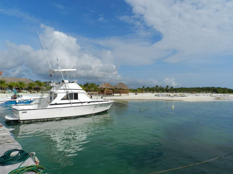 White sport-fishing boat with a tall tuna tower and outriggers moored near a dock, with a sandy beach, palapas, sun loungers and palm trees in the background under a blue sky.