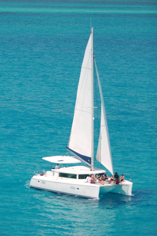 Aerial view of a white Lagoon-style sailing catamaran under white sails with a group of passengers on the foredeck, gliding over bright turquoise Caribbean water on a sunny day.