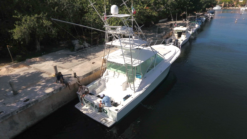 Aerial stern view of a white sportfishing yacht (transom 'OCEANO') with tuna tower moored along a concrete quay, two people sitting on the dock and crew in the cockpit. Other boats lined up behind.