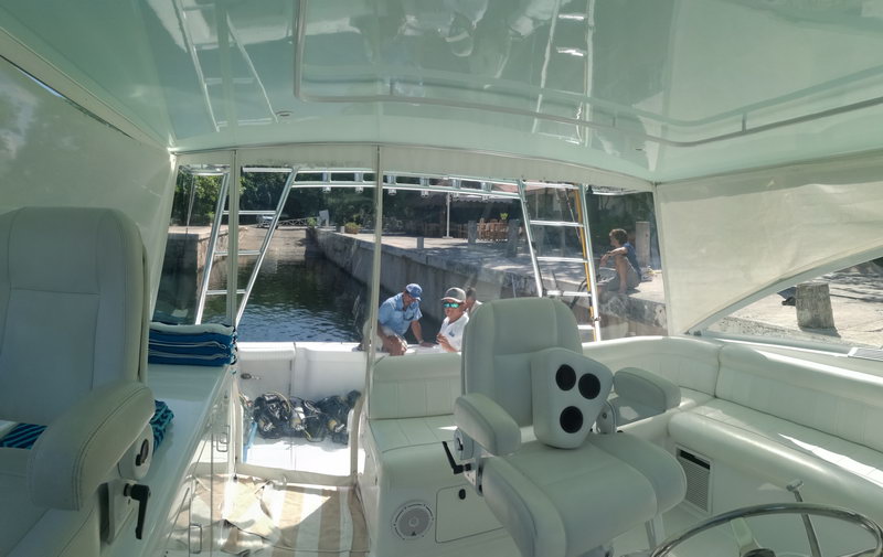 Cockpit of a sportfishing yacht looking forward, white helm seat and console with electronics, dive gear bags on the deck and two men working at the bow, dockside trees behind. Daylight.
