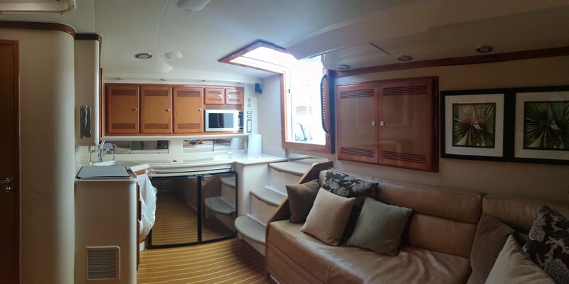 Interior of a sportfishing yacht showing the galley with wood cabinets, microwave and sink on the left, steps up to the cockpit, and a tan sofa with cushions on the right. Framed palm prints on the wall.