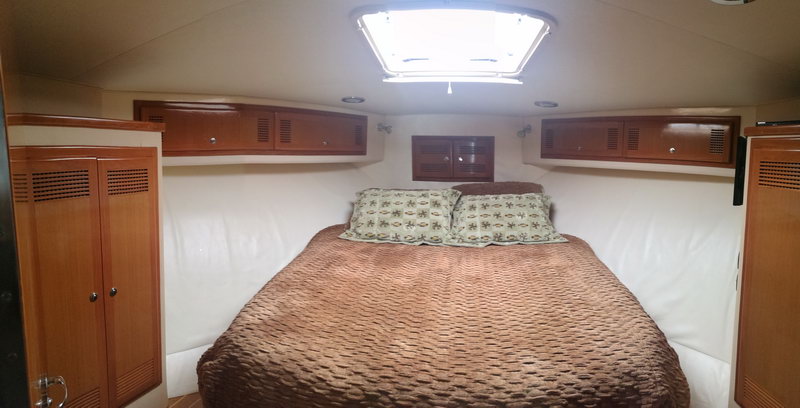 Interior master cabin of a yacht with a double bed dressed in a brown quilted cover, wood cabinetry and overhead lockers either side and an overhead hatch letting in light. Cozy enclosed berth.