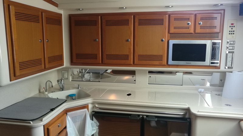 Galley of a sportfishing yacht with wood cabinet doors, a white Panasonic microwave, white countertops, sink and a control panel. Bright artificial lighting, clean modern fit-out.
