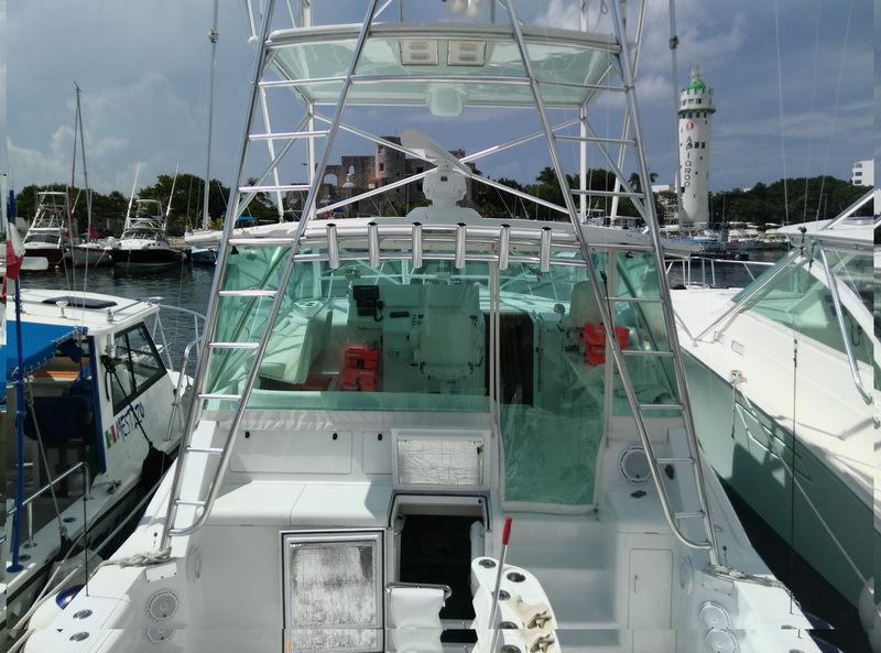Stern cockpit of a white sportfishing boat with tuna tower and clear enclosure, moored between two other boats in a Cozumel marina with a green-and-white lighthouse and stone fort behind. Sunny.