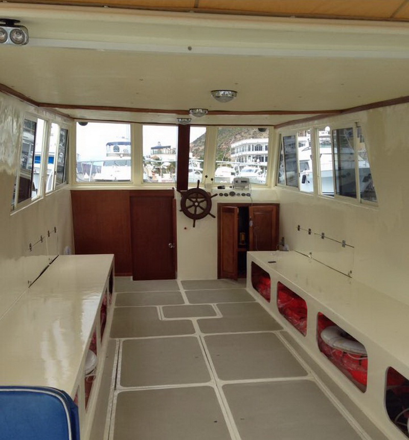 Interior main cabin of an older wooden tour boat with white-painted walls and ceiling, a wooden ship's-wheel decoration on the central bulkhead, life jackets stowed in open compartments along the sides, and marina boats visible through the windows.