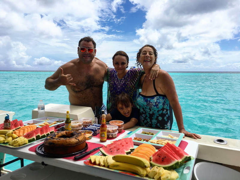 A group of adults and a child stand in turquoise water behind a boat's swim platform spread with fruit, watermelon, snacks and hot-sauce bottles, smiling and posing. Bright turquoise sea and blue sky with clouds behind them.