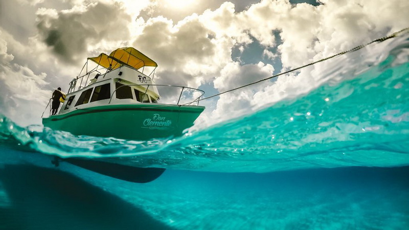 Half-over half-under split-level water shot of the green-and-white 'Don Clemente' boat with yellow canopy floating on turquoise water, dramatic puffy clouds above and clear water below. A person in yellow stands on the bow.
