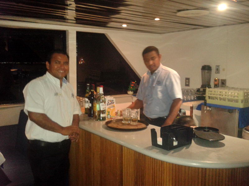 Dos cantineros sonrientes detrás de la barra libre del barco Discovery de noche preparando bebidas para los invitados