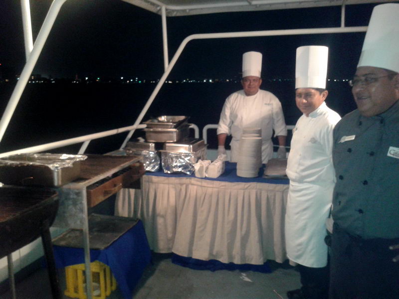 Chefs con gorros blancos en una parrilla y estación de bufé sirviendo una cena nocturna a bordo del Discovery con las luces de Cancún al fondo