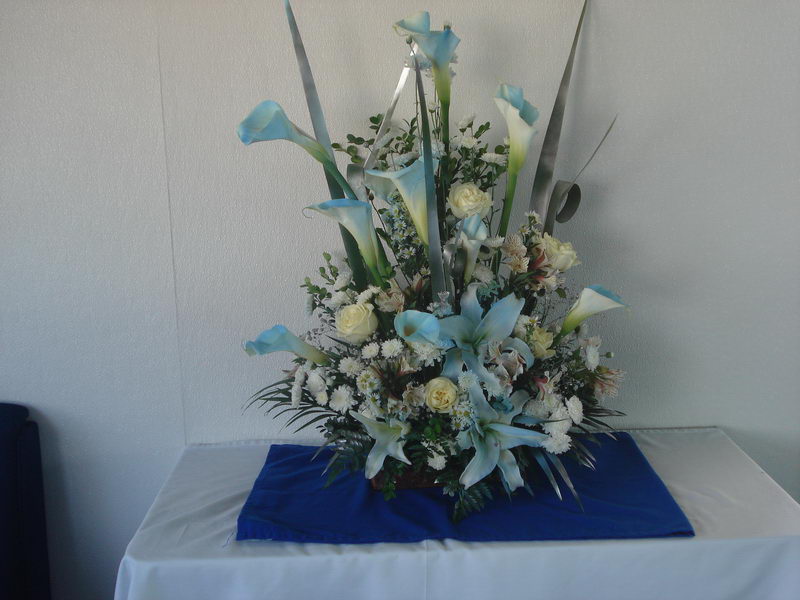 Tall floral arrangement of pale-blue calla lilies, blue lilies and cream roses against a plain white wall, sitting on a blue and white draped table.