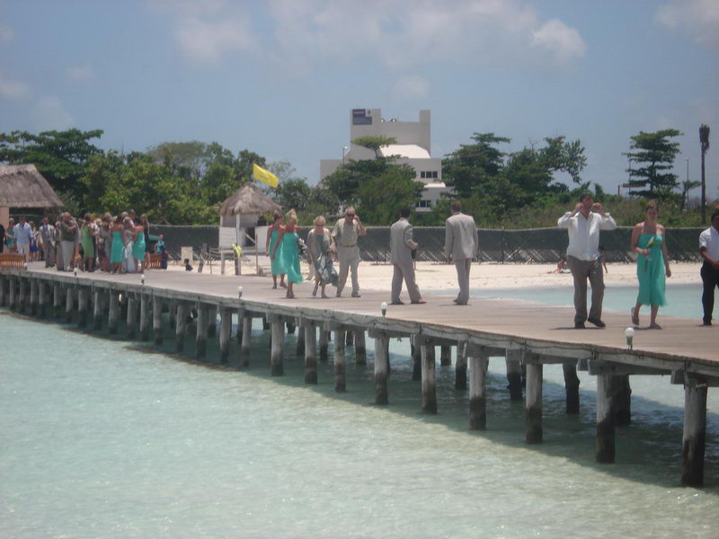 Wedding guests walking along a long wooden pier toward a sandy beach; several women in matching teal dresses, men in tan suits, turquoise shallow water, palapas and buildings ashore under a bright sky.