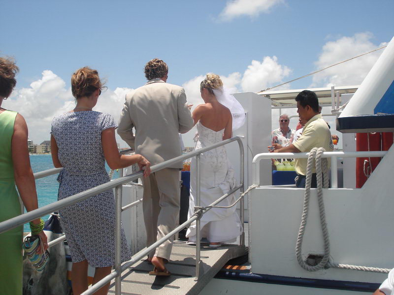 Bride in a white gown and groom in a tan suit climbing a gangway to board the boat, other guests and a crew member around them, sunny sky and sea behind, mooring rope in foreground.