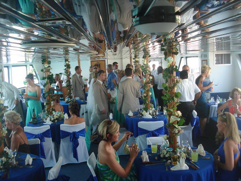 Crowded wedding reception inside the salon, guests standing and seated among ivy-wrapped columns, blue tables, women in teal, mirrored ceiling reflecting the whole scene. Bright daylight through windows.