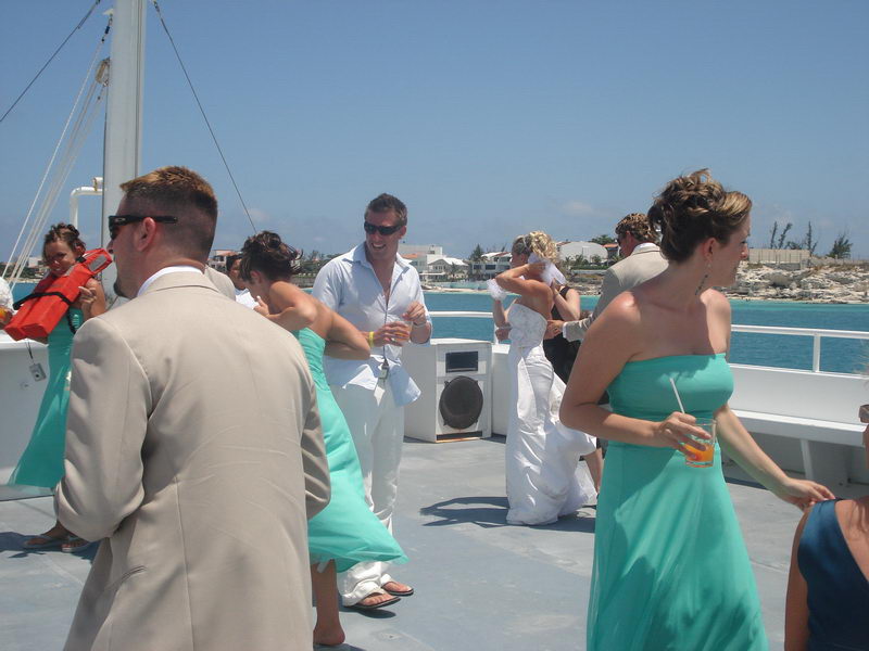 Bride and groom reclining together on the white bow deck of the boat, groom in tan suit and sunglasses, bride in white gown, deep turquoise water and blue sky behind.