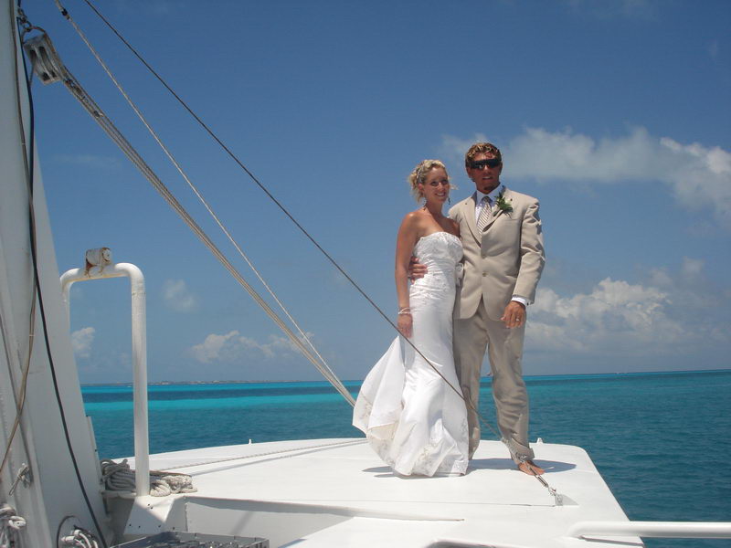 Wedding party on the open upper deck; bride and groom stand on a raised platform by the mast (Furuno radar) while guests gather below, turquoise sea, puffy clouds and bright sun.