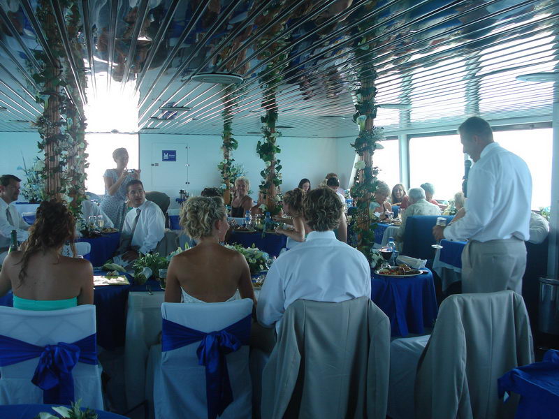 Wedding reception inside the salon, guests seated at blue tables with white-and-blue chair sashes, the groom standing with a microphone giving a toast, ivy-wrapped columns and mirrored ceiling.