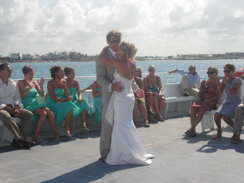 Bride and groom embracing/first-dancing on the open deck while seated guests in teal dresses look on, marina and shoreline behind under a partly cloudy sky.
