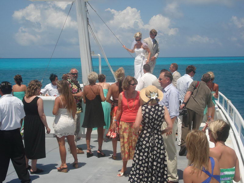 Wedding party gathered on the open deck, bride and groom standing on a raised platform, guests mingling below in summery dresses, vivid turquoise sea and blue sky behind.