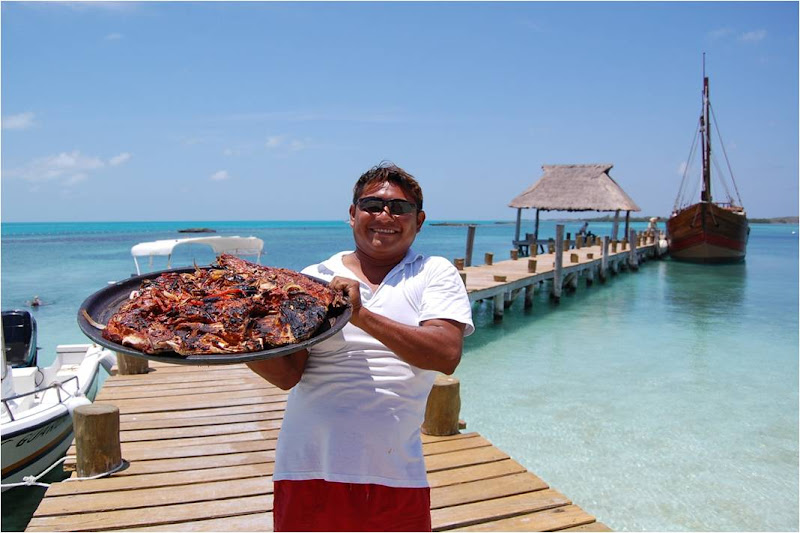 Tripulante sonriente sosteniendo una gran charola de pescado recién asado en el muelle con la carabela y el mar turquesa al fondo en Isla Contoy