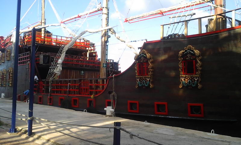 Side view of a dark wooden pirate galleon docked at a concrete pier, with red-trimmed rails, ornate gold-framed gun ports and furled sails. Rigging and masts rise above. Bright daylight.