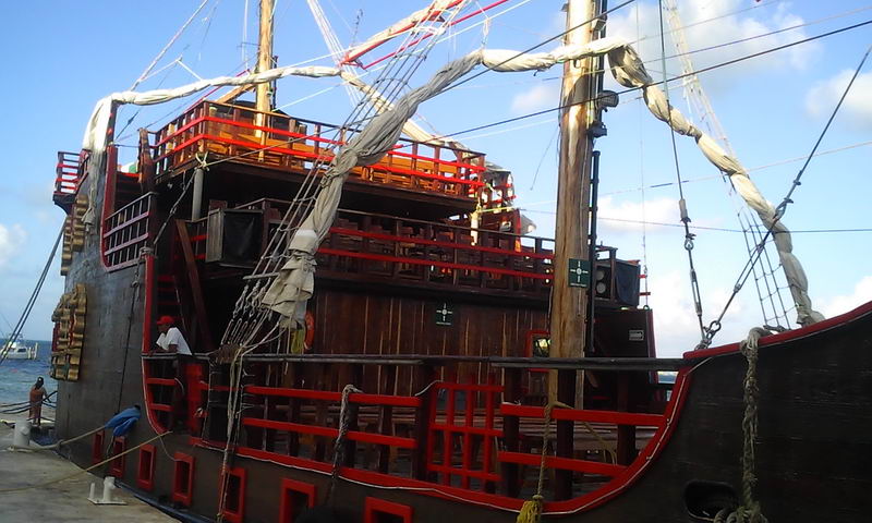 Bow view of the dark wooden pirate galleon tied at a dock, showing multiple decks with red-and-wood railings, furled white sails and heavy rope rigging. A crew member stands at the base. Clear sky.