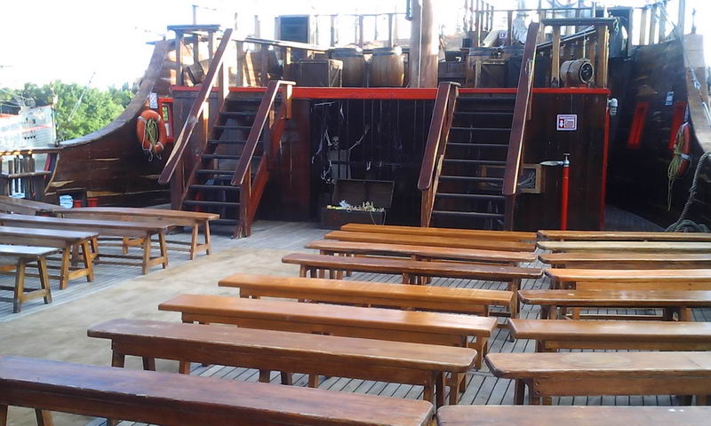 Empty main deck of the pirate galleon set up with rows of wooden bench-and-table seating for the dinner show. Staircases lead to an upper deck; life rings and a red fire extinguisher are mounted on the wood-paneled cabin. Daytime.