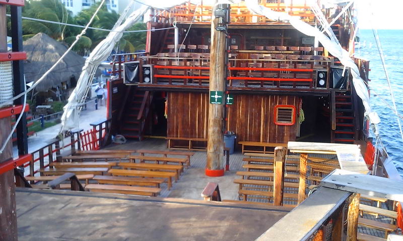 Open deck of the pirate galleon with wooden benches arranged around a central mast, ready for the show. Wood-paneled superstructure with staircases behind; the shoreline with palapas is visible to the left and open sea to the right. Daytime.