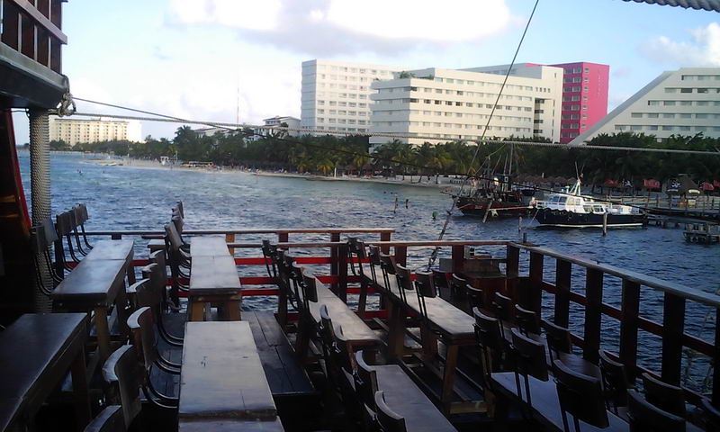 View from the deck of the Jolly Roger ship looking across the water toward the Cancun beach and large hotel buildings, with rows of wooden show benches in the foreground and other boats moored nearby. Overcast late-day light.