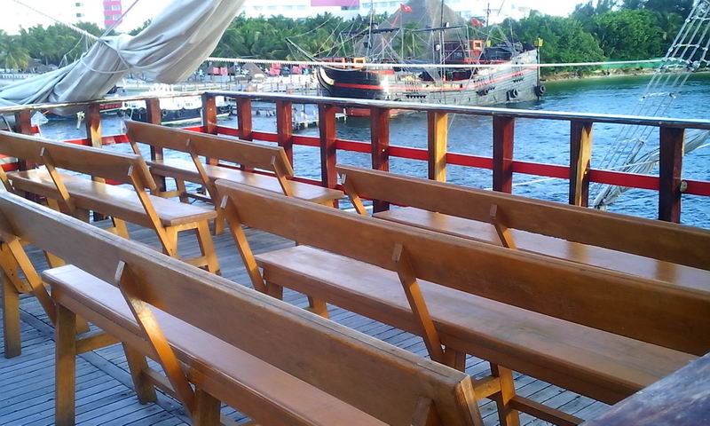 Rows of varnished wooden bench seats on the deck of one pirate ship, looking across the water toward a second dark pirate galleon moored near a palm-lined shore in the Cancun hotel zone. Soft evening light.