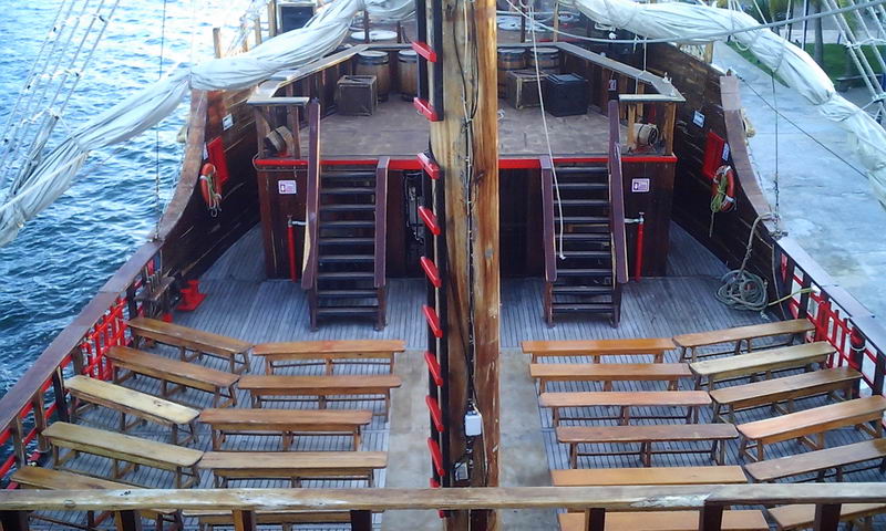 Overhead view of the pirate galleon's lower deck showing symmetrical rows of wooden benches, two staircases leading up, a central mast and furled sails along the rails. Open water on the left. Daytime.