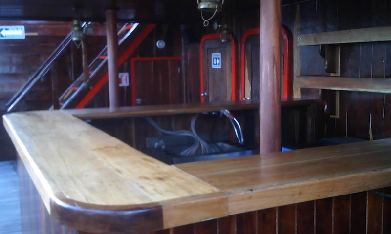 Interior wooden bar on the pirate ship with a polished wood countertop and paneled walls, a staircase and red emergency-exit signage behind. Dim indoor lighting; some wiring and equipment visible below the bar.
