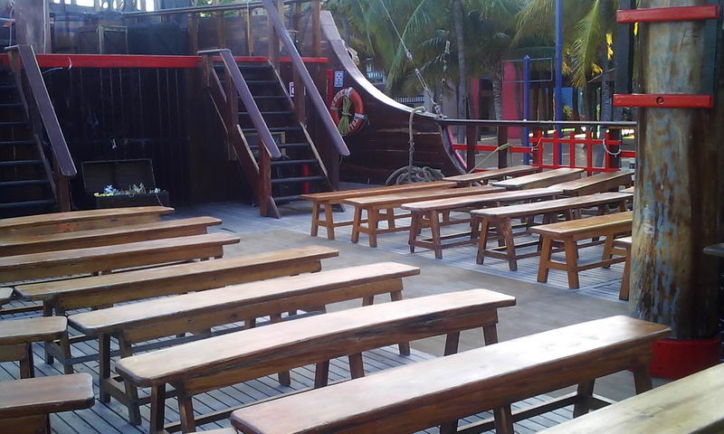 Deck of the pirate galleon with rows of wooden benches and tables set for the show, staircases and a treasure chest at center, palm trees and a colorful playground structure visible on shore behind. Bright daytime.