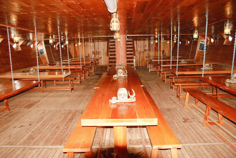 Interior lower dining deck of the pirate ship with long varnished wooden tables and benches lit by hanging oil-style lanterns, decorative skulls placed along the tables, and a central staircase. Warm wood tones throughout.