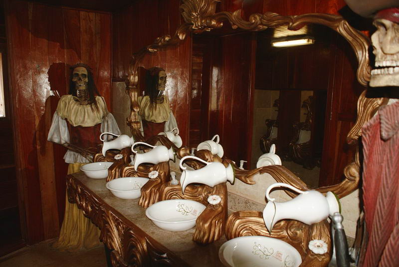 Themed restroom on the pirate ship with white porcelain pitcher-and-bowl washbasins on ornate gilded stands, wood-paneled walls, large mirror, and a costumed female skeleton mannequin in period dress standing beside them.