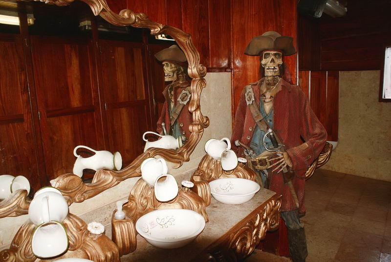 Themed washroom aboard the pirate ship with white pitcher-and-bowl basins on gilded pedestals, a large ornate mirror, wood paneling, and a skeleton mannequin dressed as a pirate in a red coat and tricorn hat standing at right.